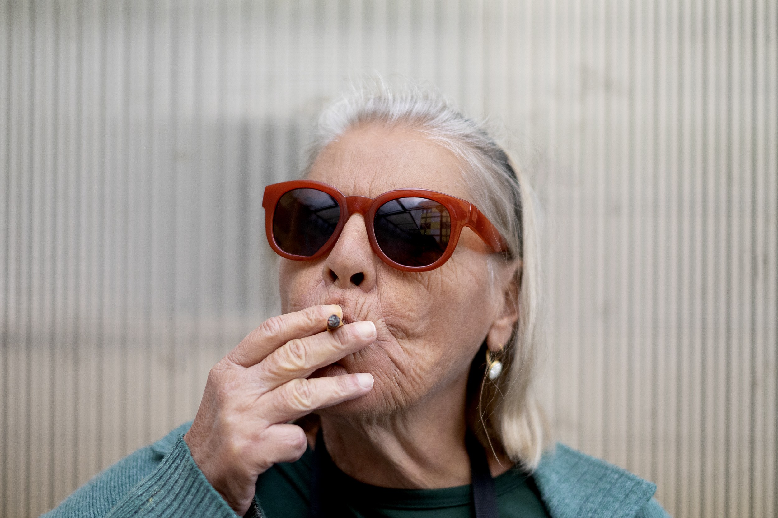 Mulher idosa com óculos escuros de armação vermelha fuma cigarro com a mão direita.