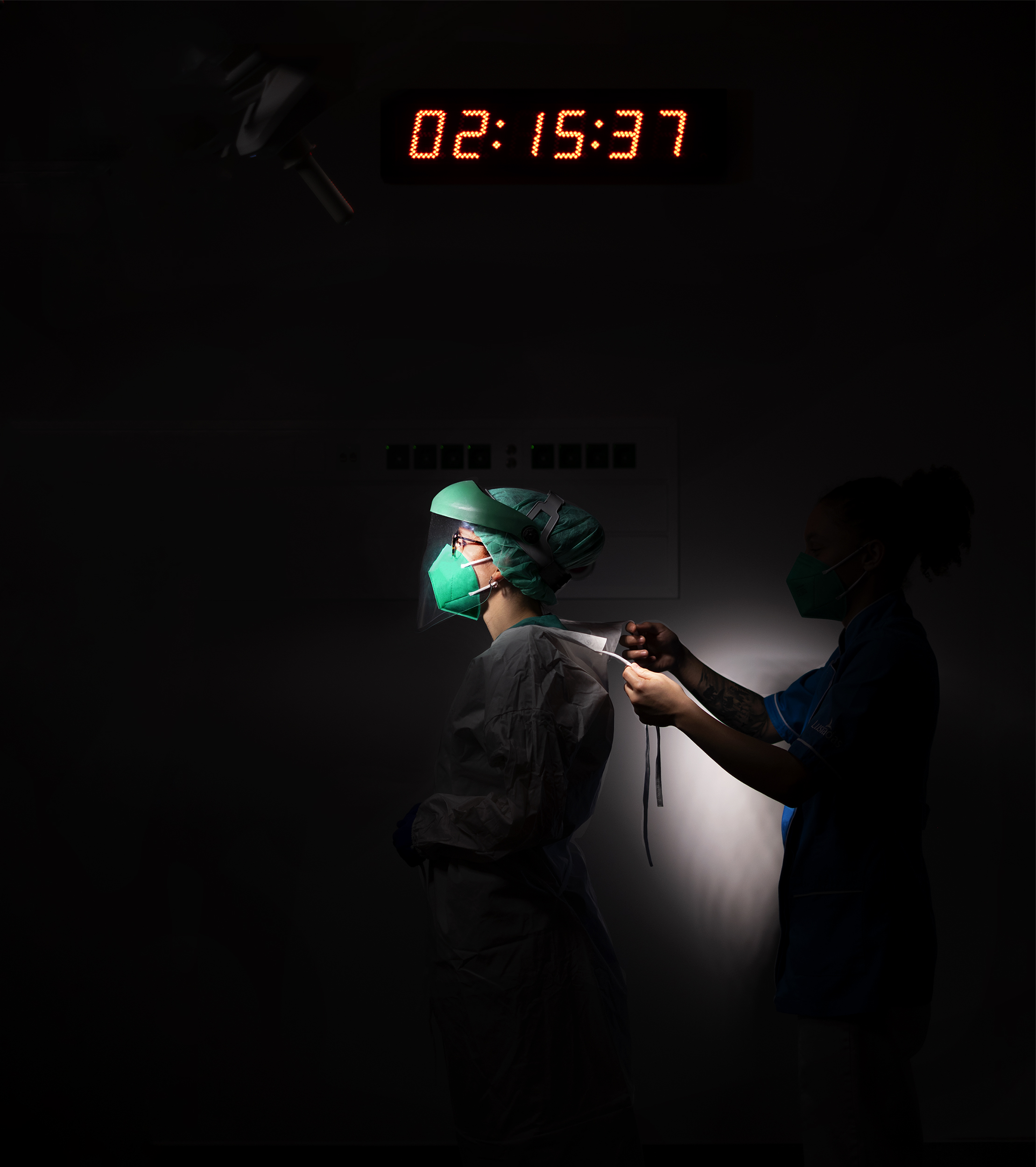 Enfermeira de perfil em ambiente escuro ajudada por outra enfermeira a colocar equipamento de proteção individual.