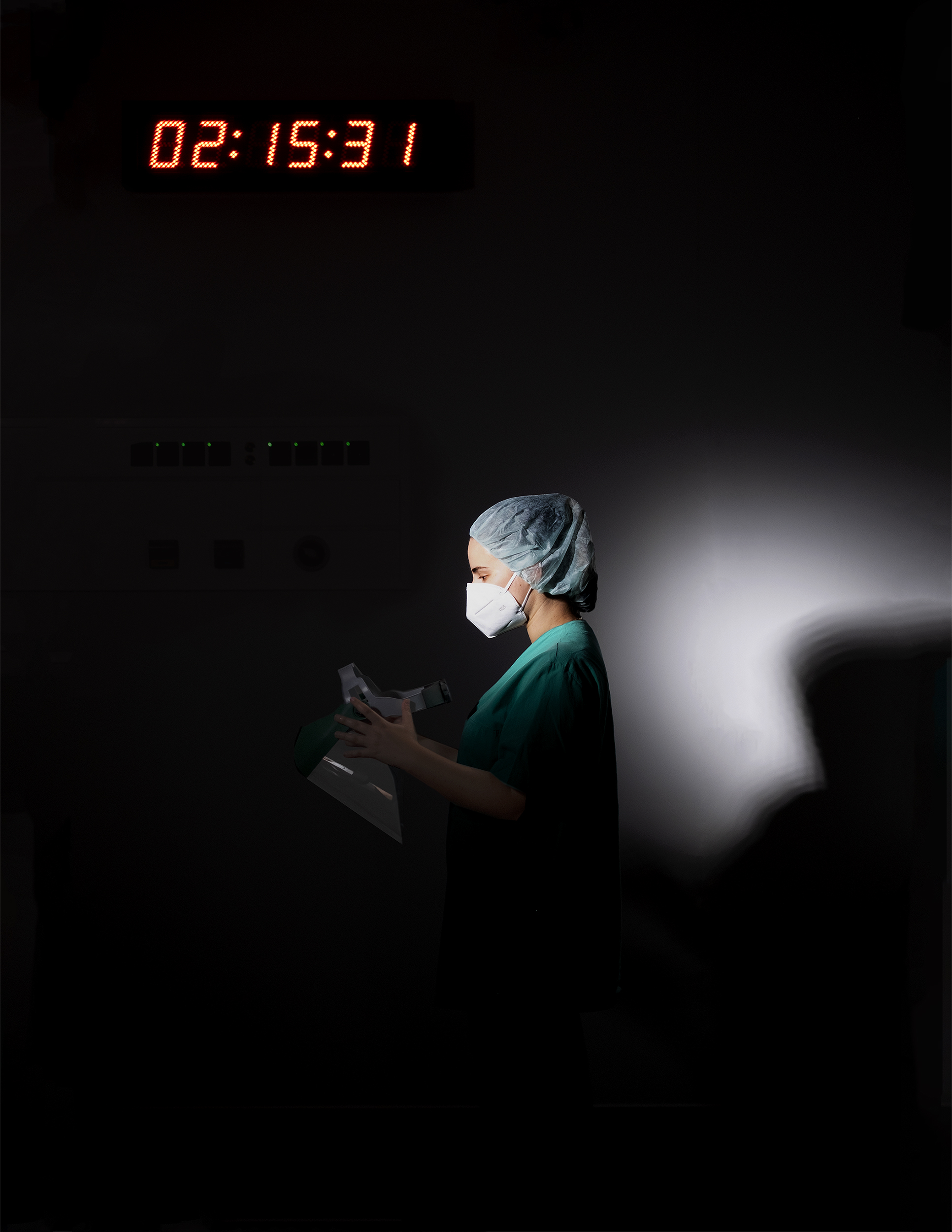 Enfermeira de perfil em ambiente escuro com máscara e touca, segura viseira.