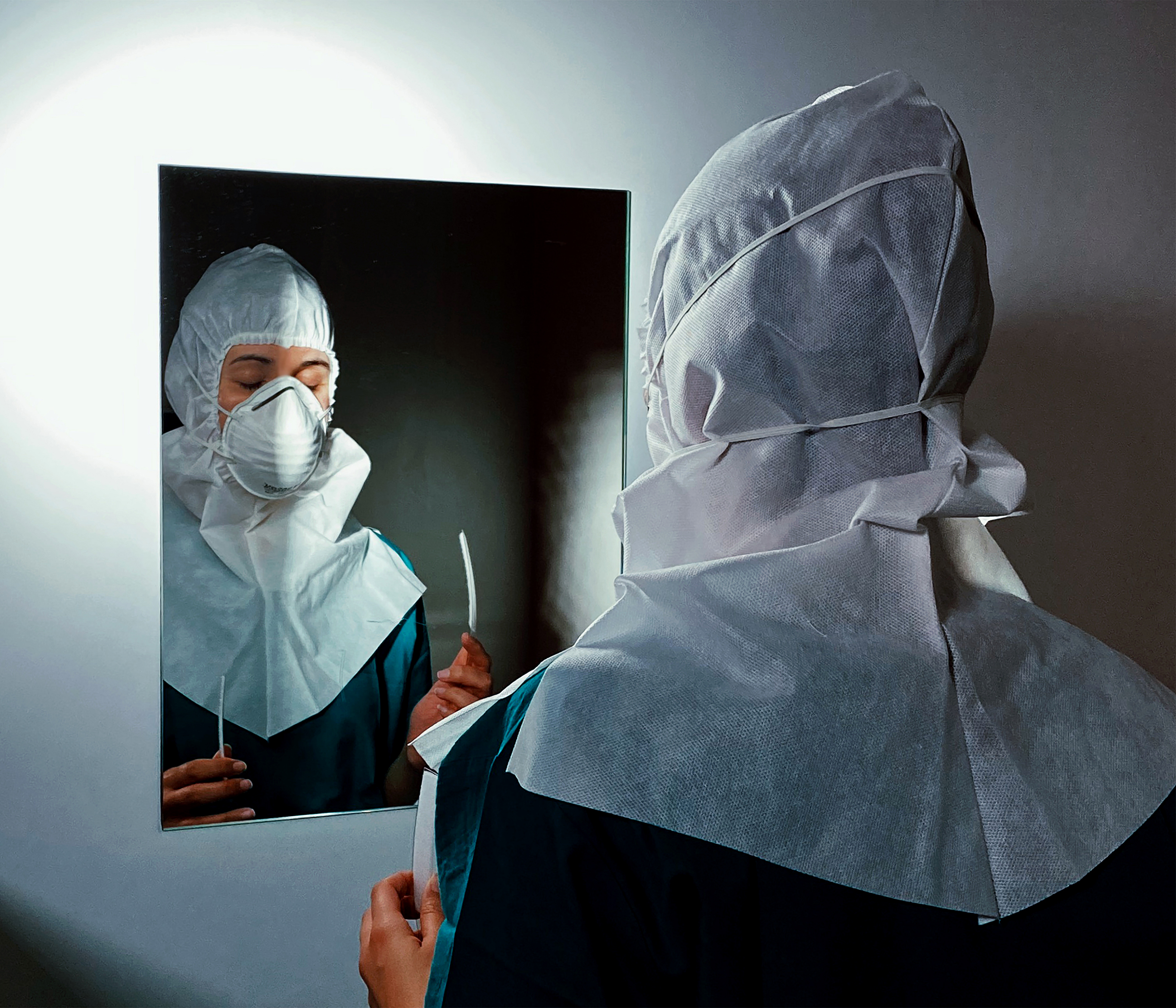 Enfermeira de olhos fechados usando máscara e capuz, refletida em espelho, coloca viseira.