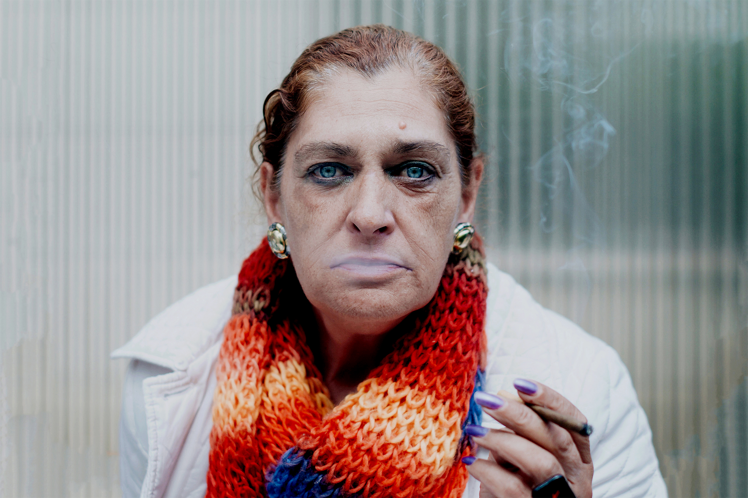 Mulher de olhos azuis encara a Câmara, usa cachecol vermelho e fuma cigarro.